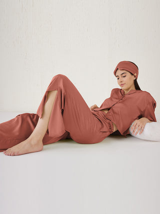 A woman in a black silk pajama set and sleep mask poses with her hands behind her head.