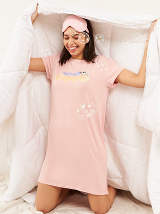 A woman stands on a bed, wearing a pink, short-sleeve sleep dress with a "he's all good" graphic and a pink eye mask.